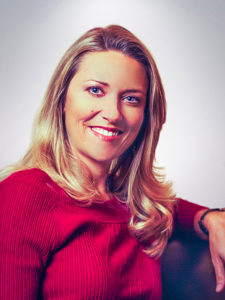 A smiling woman with long blonde hair wearing a red sweater sits in front of a neutral background, looking towards the camera.