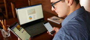 A person in a blue shirt, donning glasses, deftly juggles a tablet and smartphone at their desk. A laptop before them displays Account Based Marketing graphs and charts, while a cup of pencils stands ready nearby.