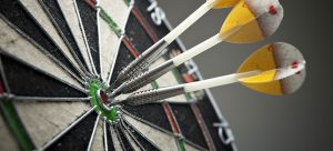Close-up of three darts hitting the bullseye on a dartboard, much like Account Based Marketing targets specific prospects. The darts, with yellow and white flights and metal shafts, demonstrate precision and accuracy. The background is blurred, emphasizing the focus on the dartboard.