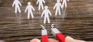 A hand holding a red horseshoe magnet is irresistibly attracting paper cutouts of people on a wooden surface, symbolizing the genuine power of attracting talent or customers without false promises.