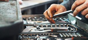 Hands selecting a tool from a large toolkit filled with various wrenches, sockets, and screwdrivers. The tools are organized neatly in a portable toolbox, suggesting a work setting like a garage or workshop.