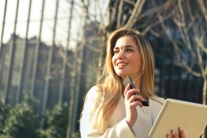 A woman with long hair smiles while holding a smartphone and a tablet. She is wearing a white coat and is standing outdoors, with trees and a building in the background. The sunlight is casting a warm glow on her face.