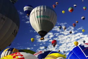 A vibrant sky filled with colorful hot air balloons of various sizes, floating under a blue sky with scattered white clouds. The balloons display diverse patterns and logos, creating a lively scene.