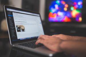 A person typing on a laptop displaying a blogging platform. The screen shows a blog post with an image of a latte. A colorful, blurred television screen is visible in the background, suggesting a cozy or creative workspace.
