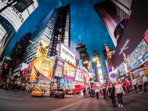 A vibrant nighttime scene at Times Square featuring illuminated billboards, bustling crowds, taxis, and the iconic architecture of New York City. The sky is a deep blue, enhancing the colorful lights and busy atmosphere of the area.