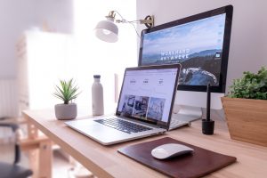A modern home office setup featuring a laptop and desktop computer on a wooden desk. The laptop displays graphs, while the desktop screen has the text "Work Hard Anywhere" over a scenic background. A plant, a water bottle, and a lamp are also on the desk.