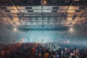 A large indoor concert with a lively crowd. The stage is brightly lit, and the performer stands at the center, raising their arms. The audience is enthusiastically reaching towards the stage, creating an energetic atmosphere.