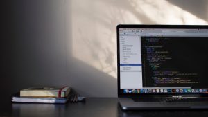 A laptop displaying code sits on a desk next to a stack of books and some pencils. Sunlight casts shadows on the wall, creating a calm atmosphere. The text editor on the screen shows colorful syntax highlighting.