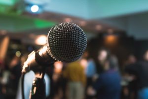 Close-up of a microphone on a stand, with a blurred audience and dimly lit room in the background, suggesting a live performance or public speaking event.