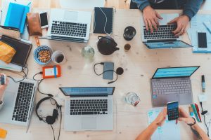 Aerial view of a wooden table with four laptops, notebooks, phones, and other office items. Several people are working, using laptops and smartphones. The table is cluttered with tech gadgets and snacks, including a bowl of cereal and a teapot.
