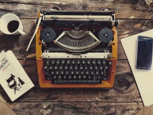 A vintage typewriter sits on a rustic wooden table surrounded by a cup of coffee, a book, a notepad, and a smartphone.