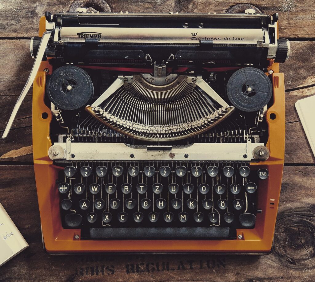 A vintage typewriter sits on a rustic wooden table surrounded by a cup of coffee, a book, a notepad, and a smartphone.
