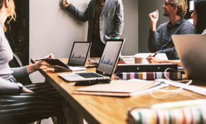A group of people in a meeting room focuses on account-based marketing strategies, with laptops displaying charts open on a wooden table. One person stands at a whiteboard, gesturing, while others sit and listen attentively. Books and coffee cups are also on the table.