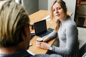 A woman with long hair, wearing a gray sweater, sits at a wooden table holding a cup. A laptop is open in front of her as she engages in conversation with someone whose back is to the camera. A phone rests on the table, capturing this seemingly candid moment that feels far from false.