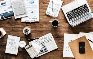 A wooden desk with scattered work items: a laptop, coffee cups, a smartphone falsely displaying 08:15, a calculator, business newspapers, charts, papers, and potted plants. The setup suggests a busy yet organized workspace.