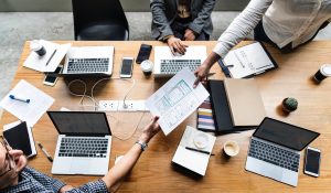 A wooden table with six laptops, notebooks, coffee cups, and a small cactus. Four people are seated, with one handing a document to another. A power strip lies on the table, connecting the devices. Collaborative workspace setting.