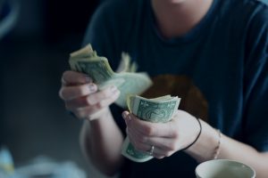 A person in a blue shirt is holding and counting a seemingly false stack of U.S. one-dollar bills, with a focus on the hands and money. A portion of a coffee cup is visible in the blurred background's lower right corner.