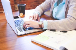 A person wearing a watch is typing on a laptop at a wooden table. A notebook with handwritten notes and a pen are nearby. There's also a black coffee cup on the left side of the table.
