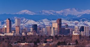 A cityscape view of Denver, Colorado, at dusk. Skyscrapers rise against a backdrop of snow-capped mountains under a clear sky. The foreground features bare trees, reflecting the winter season, while hints of modern progress echo the energy akin to a thriving Denver SEO agency.