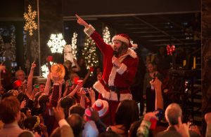 Dressed as Santa Claus, a person enthusiastically addresses a lively crowd at a festive indoor event. The crowd holds up red cups in celebration, with snowflake lights twinkling overhead—perfect inspiration for how to plan a digital marketing campaign that captures such cheerful holiday vibes.