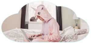 A person in a pink unicorn onesie types on a laptop, likely exploring B2B digital marketing trends, while sitting on a bed. In the background, a cat perches on the window sill. The whimsical scene is framed in a cloud shape.