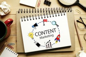 A spiral notebook on a desk displays "Content Marketing" alongside 2019 Content Trends icons: a light bulb, target, and rocket. Nearby are a coffee cup, crumpled paper, paperclips, and magnifying glass.