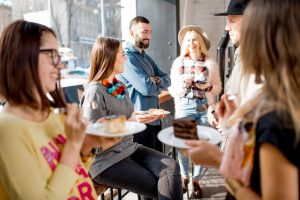 A group of people socializing indoors, holding plates of cake and glasses, enjoying the benefits of employee advocacy. They are smiling and engaged in conversation, wearing casual clothes. The background features a window with sunlight streaming in.