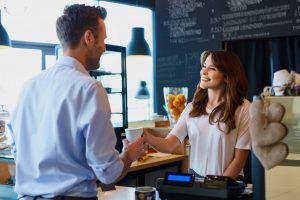 A woman in a white shirt stands behind a counter, sharing the benefits of employee advocacy as she hands a cup to a man in a blue shirt. The scene is warmed by the chalkboard menu, baked goods display, and sleek black pendant lights.