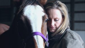 A woman with blonde hair leans affectionately against a horse with a white blaze, wearing a purple halter. The setting appears to be indoors, with soft lighting creating an intimate atmosphere.
