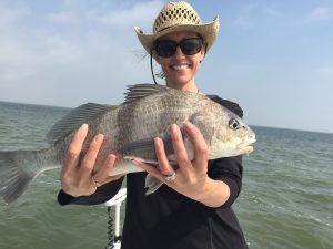 Person wearing a straw hat and sunglasses, smiling, while holding a large fish on a boat. The background shows a calm body of water under a clear sky.