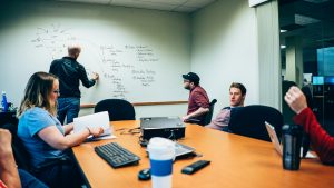 A group of people in a conference room are engaged in a discussion. One person is writing on a whiteboard filled with diagrams and notes, while others sit around a table with laptops and notebooks. There are various beverages on the table.