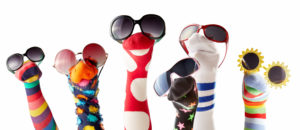 A group of colorful sock puppets wearing sunglasses. Each sock has a different design and pattern, including stripes, polka dots, and stars, with some featuring fuzzy textures. The puppets are arranged in a playful row against a white background.
