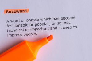 A close-up image of the definition of "buzzword" printed on paper, highlighted with an orange marker. The definition explains it as a word or phrase that is fashionable or technical, used to impress people. An uncapped orange highlighter lies on the paper.