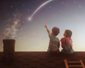 A boy and girl sit on a rooftop at dusk, gazing at a shooting star in the sky. The boy points towards the star. A chimney is visible to the left, and a small wooden ladder is on the right.