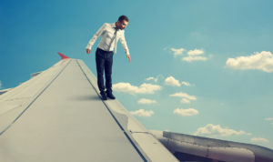 A man in business attire, embodying the spirit of agile marketing, stands on the wing of an airplane in flight under a clear blue sky with scattered clouds, looking down with concern. The scene conveys a surreal and daring situation.