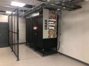 A room with a black server cabinet labeled "GRAH" next to a colorful panel with artwork. The space has gray flooring, a metal mesh partition, and cable trays on the ceiling.