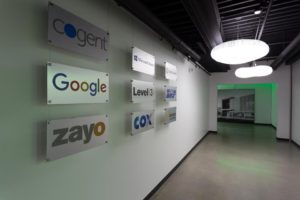 A hallway featuring illuminated signs with logos of technology and telecommunications companies, including Cogent, Zayo, Google, Microsoft Azure, Level(3), and Cox, on a white wall. Modern ceiling lights are visible above the space.