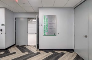 A modern hallway with gray walls and a chevron-patterned floor leads to an open door marked "Customer Storage & Operations." A sign on the wall lists directions and rooms such as "Operations Center" and "Office Suites.