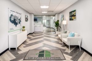A modern office lobby with white walls and a chevron-patterned floor. It has two white chairs with pillows, a green plant, and framed pictures. A map is on the wall and a mat on the floor reads "H5 DATA CENTERS.