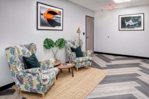 A cozy room with two patterned armchairs, a small side table, and green cushions. Two framed photos hang on the walls. A large plant and a woven rug add warmth. The floor has a chevron pattern, and a door is visible in the background.
