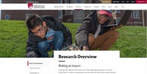 Two students work outdoors on a grassy slope at the University of Denver. One student examines the ground, while the other holds a laptop. The university's logo is visible at the top of the image, alongside navigation options for their website.