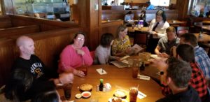A group of people gather around a large wooden table in a restaurant, engaged in conversation. Drinks and bowls of food are on the table. A server in the background is taking an order. The atmosphere is casual and lively.