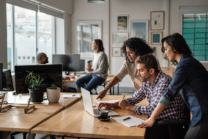 A group of professionals collaborate in a modern office. Three people focus on a laptop, while a woman sits casually on a desk in the background holding a coffee cup. The space is filled with natural light and potted plants.