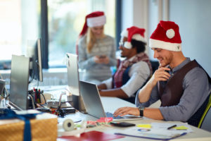 Office workers wearing Santa hats sit at their desks, using computers. A man in the foreground smiles at his laptop. In the background, a woman checks her phone, and another person chats. A wrapped gift is on the table. It's a festive setting.