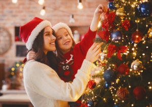 A woman and a child, both wearing Santa hats, decorate a Christmas tree with ornaments and lights. The scene is warmly lit, creating a cozy holiday atmosphere. The child smiles while reaching up to place an ornament.