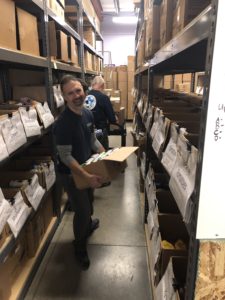 A man in a dark shirt smiles and carries a box down an aisle filled with shelves of organized food items. The shelves are lined with labeled cardboard boxes. Another person is visible in the background, also working.