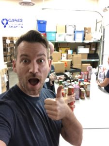 A man taking a selfie in a storage room with shelves and boxes. He is giving a thumbs-up and smiling with an open mouth. Two people are organizing items on a table behind him, with various packaged goods visible.