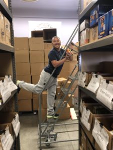 A person smiles while balancing on one leg on a ladder amidst stacked boxes in a storage area. They are wearing a navy shirt, gray sweatpants, and holding a tape dispenser. Shelves on both sides are labeled with different food items.
