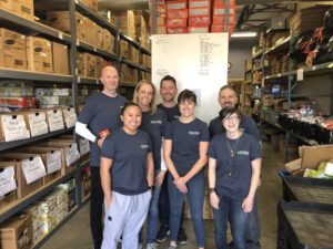 A group of seven people in matching gray t-shirts stands in a warehouse filled with boxes and shelves. They are smiling and appear to be volunteering, possibly organizing food or supplies.