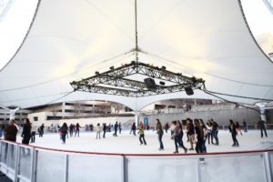 People ice skating under a large, white, tent-like canopy. The rink is surrounded by transparent barriers, and several skaters are visible, enjoying the activity. The background includes buildings and trees.